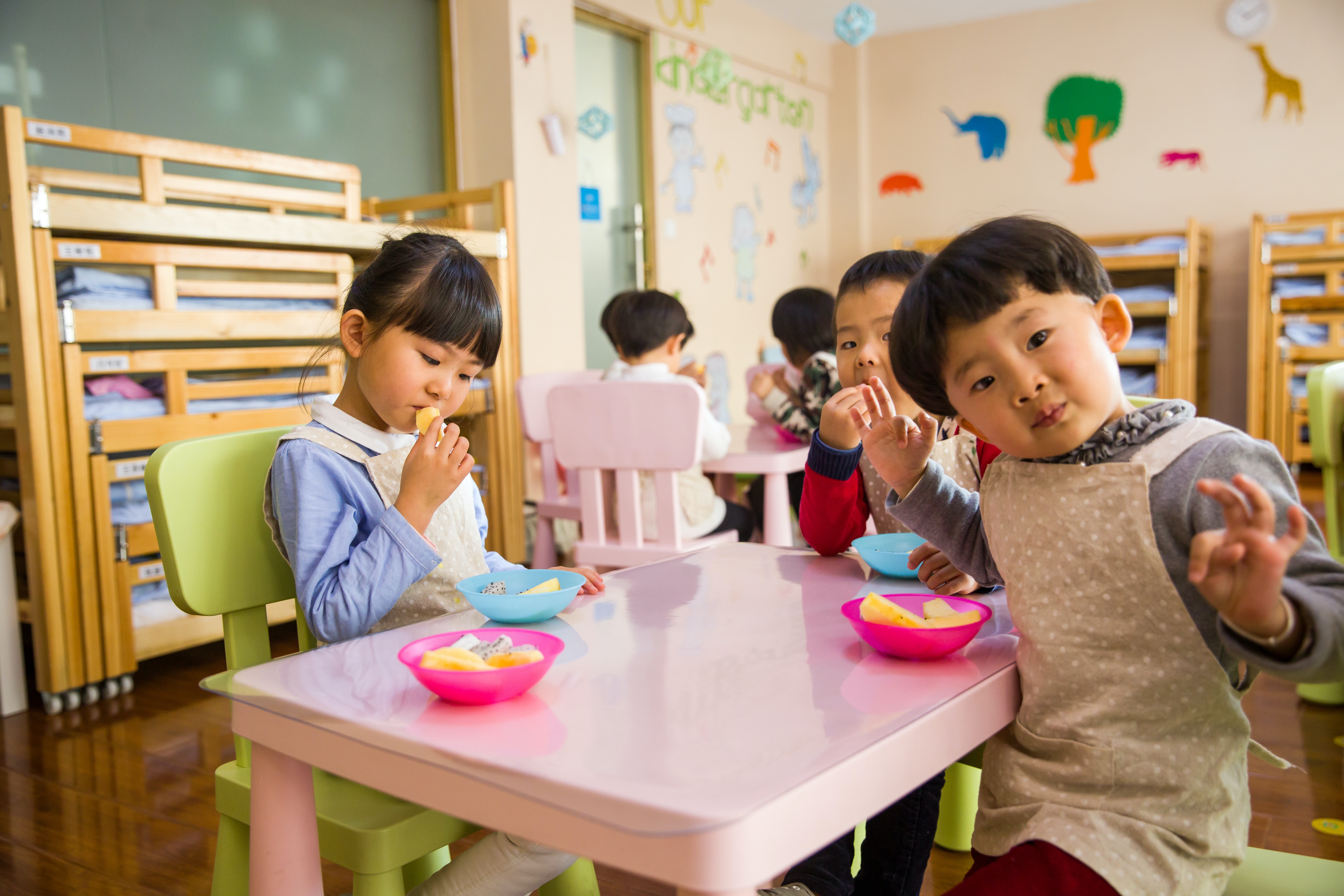 Group of young children eating snacks at a pink table, with one child in the foreground wearing an apron and looking playfully at the camera. Bunk beds are visible in the background.