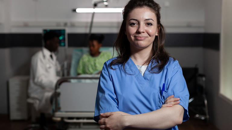 Confident nurse in blue scrubs standing with arms crossed in a hospital room, with a doctor and patient in the background.