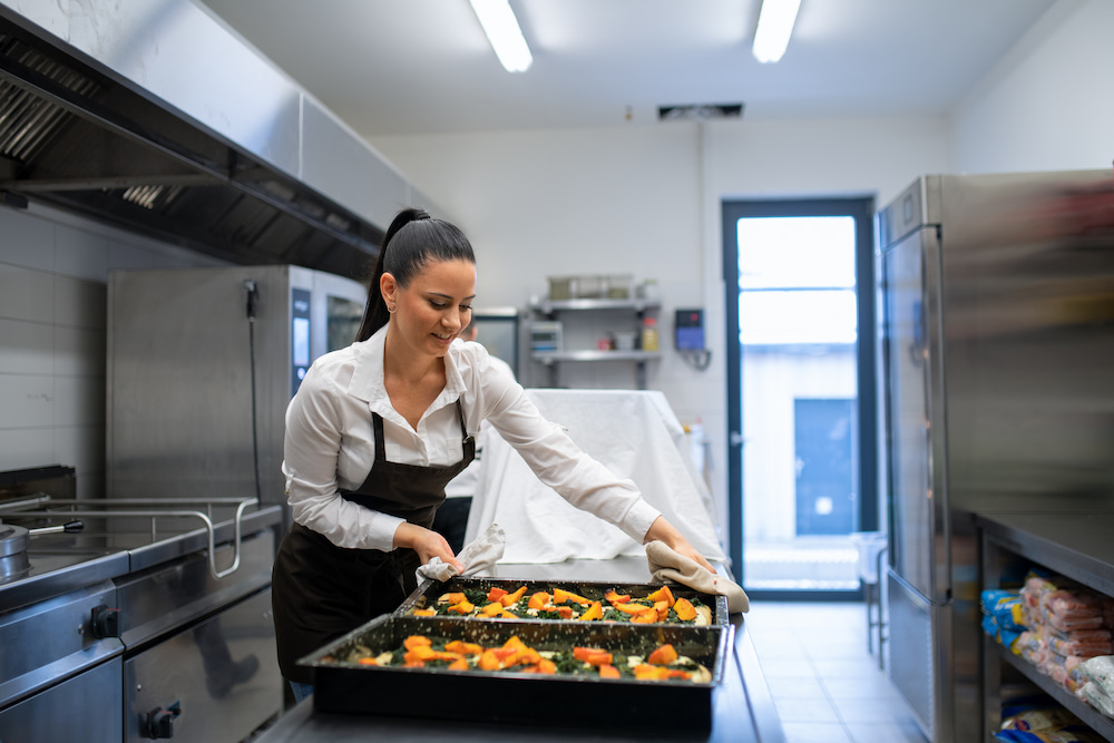 Smiling woman in a white shirt and black apron placing a large black baking tray with baked vegetables onto a metal counter in a well-lit commercial kitchen, using oven mitts.