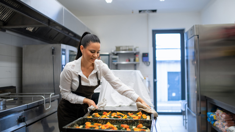 Smiling woman in a white shirt and black apron placing a large black baking tray with baked vegetables onto a metal counter in a well-lit commercial kitchen, using oven mitts.
