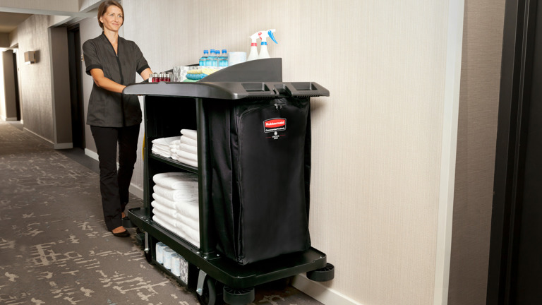 A hotel housekeeper s stands beside a Rubbermaid's Executive Traditional Full-Size Housekeeping Cart filled with towels and cleaning equipment