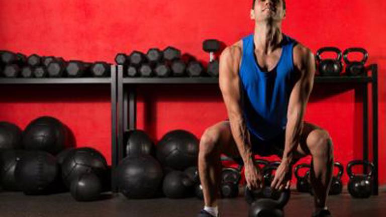 Muscular man in a blue tank top and black shorts performing a squat with kettlebells in a gym, with red wall and racks of weights in the background.