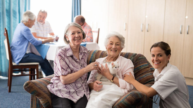 Three happy women, two elderly and one younger caregiver, sit with a small kitten in a communal living area.