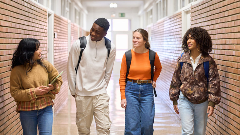 Four young adult students walking side by side in a school hallway, chatting and smiling with backpacks on.