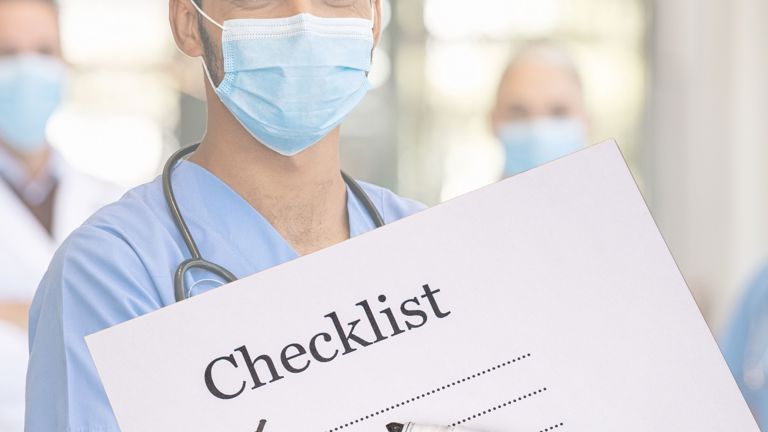 Male healthcare professional in blue scrubs and face mask holding a checklist with first four items ticked, stethoscope around his neck, in a bright hospital setting.